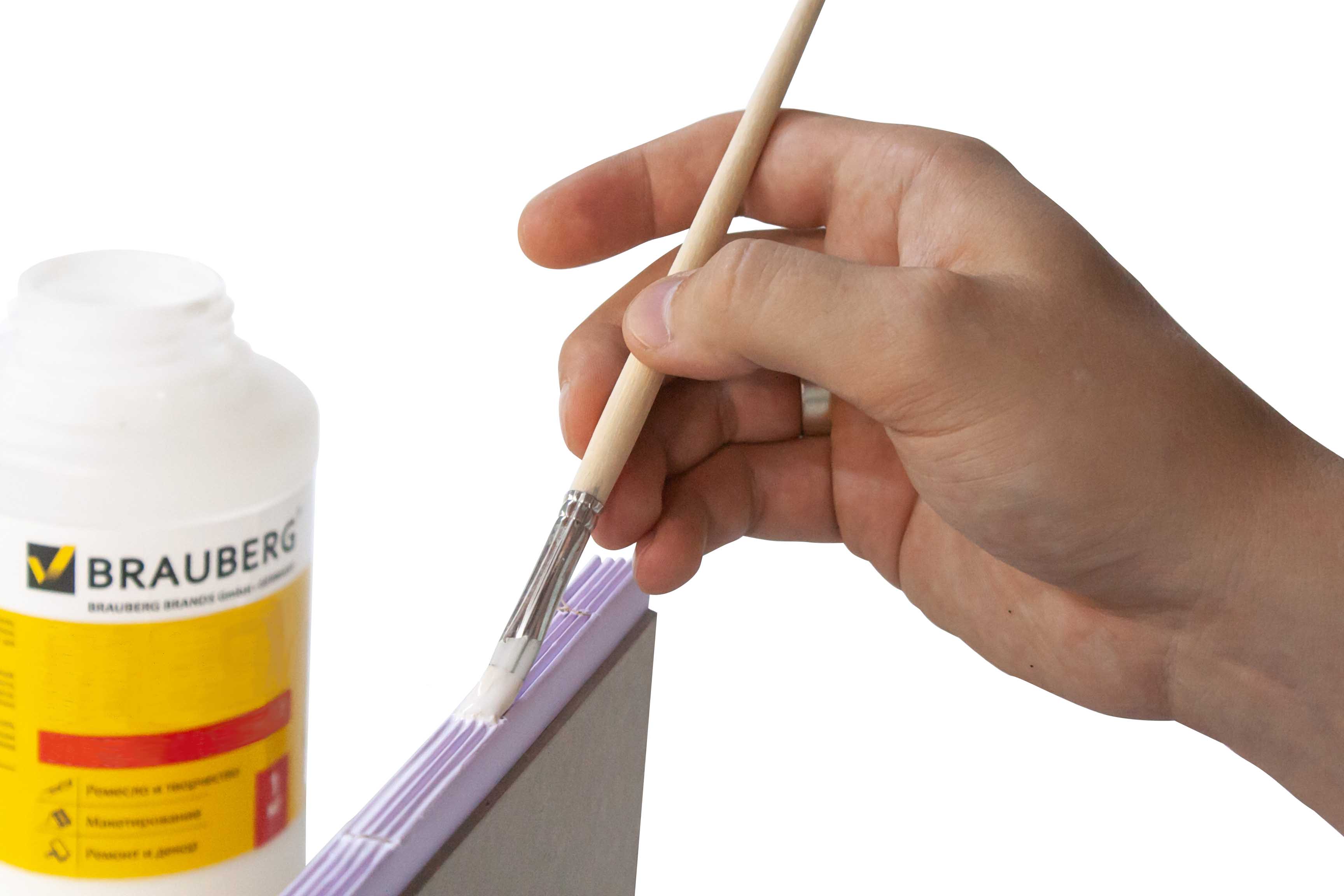 Bookbinding. A hand applying glue with a brush to the edge of a stack of paper sheets, with a glue container in the background.
