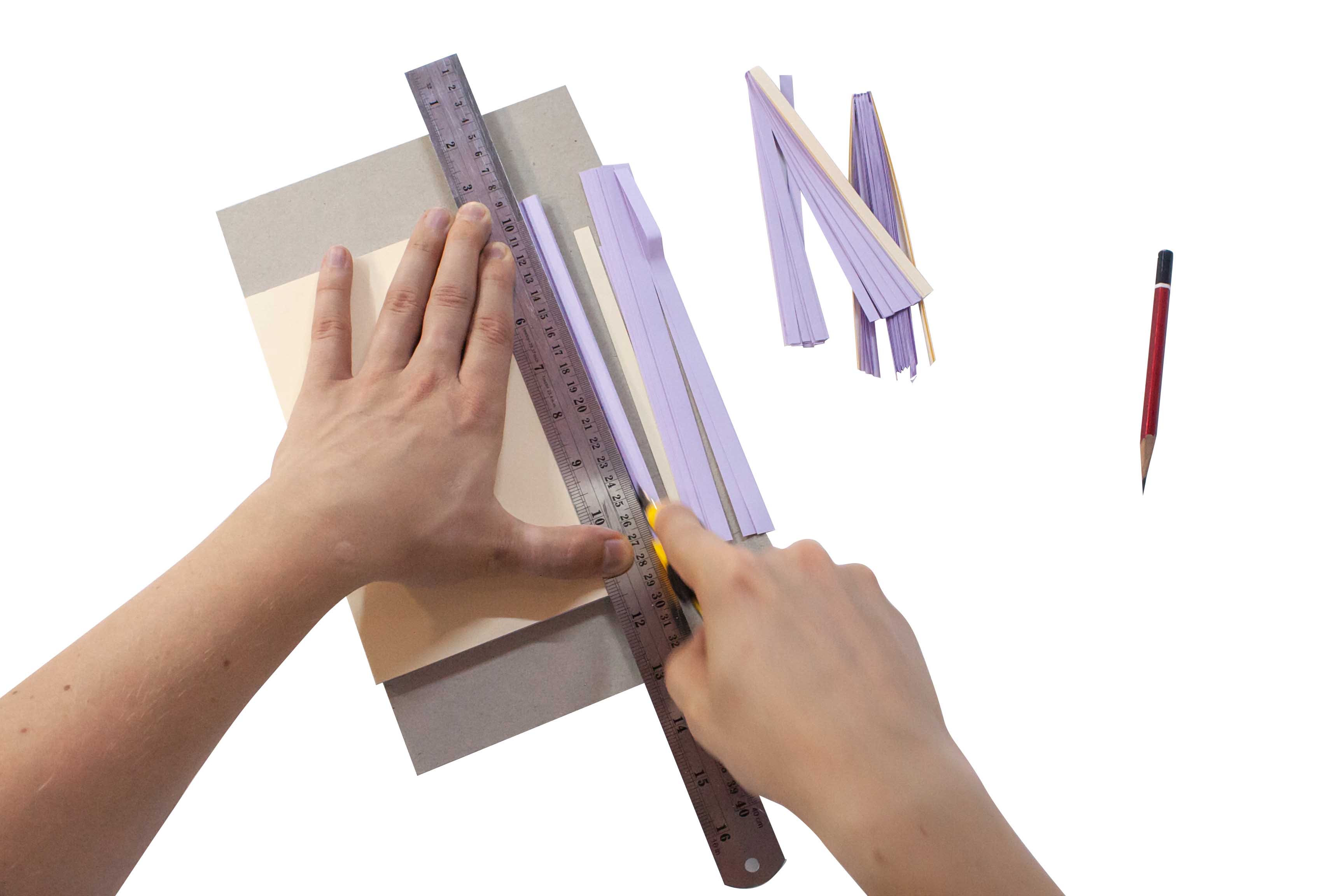 A person's hands using a ruler and a utility knife to cut along a straight edge on a piece of paper, with strips of cut paper and a pencil nearby.